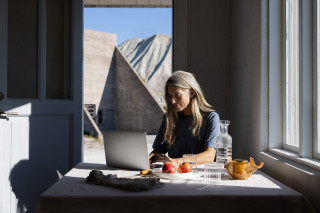 Person working on a laptop at a table near a window, photograph.